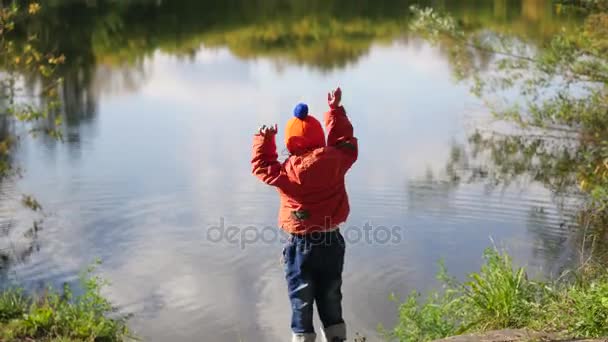 Dítě stojí na břehu rybníka a házení kamenů. Procházky na čerstvém vzduchu - Záběry, video