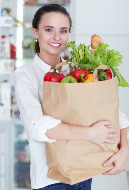 Young woman holding grocery shopping bag with vegetables .Standing in the kitchen. Woman in the kitchen looking at the camera - Fotografie, Obrázek