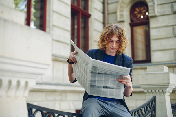 Young reddish man reading newspaper near old style building - Foto, afbeelding