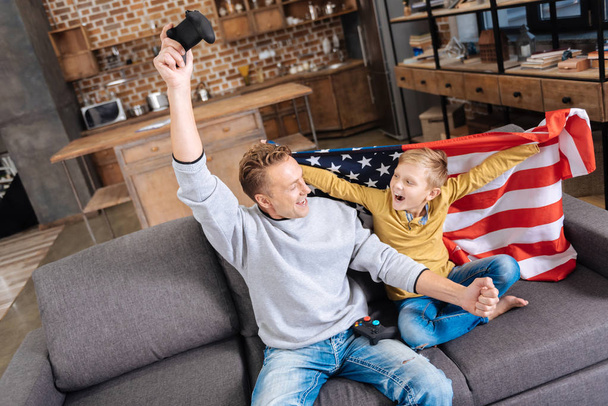 Cheerful boy wrapped in US flag cheering with dad - 写真・画像