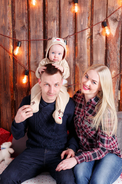 Счастливая семья на Рождество. Родители и ребенок сидят. Стена из деревянных досок и гирлянд
 - Фото, изображение