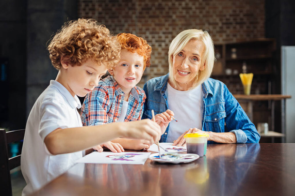 Kind elderly lady painting with grandsons at table - Foto, imagen