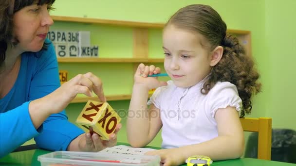 Beautiful little schoolgirl with a beautiful hairdo and a beautiful smile in a white blouse sits at her desk with a teacher and teaches foreign languages, performs a teacher's task to correctly speak and pronounce words to know the pronunciation and  - Filmati, video