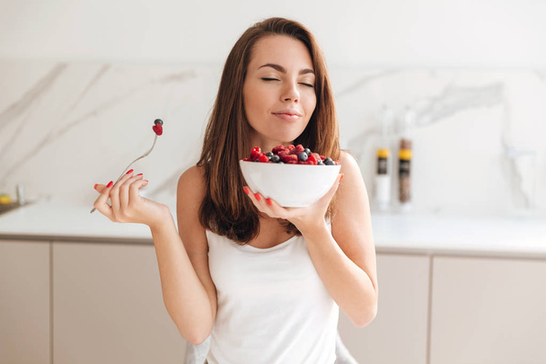 Pretty young woman enjoys eating fresh berries from a bowl - Фото, изображение