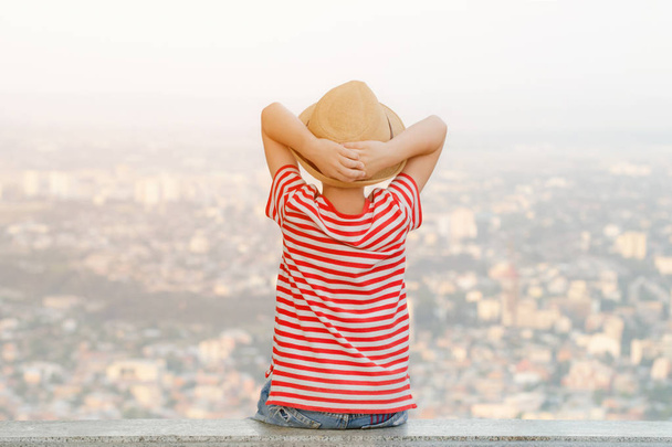 Bir şapkalı çocuk ellerini kafasının arkasında oturur ve city adlı bir yükseklikten görünüyor. Arkadan görünüş - Fotoğraf, Görsel