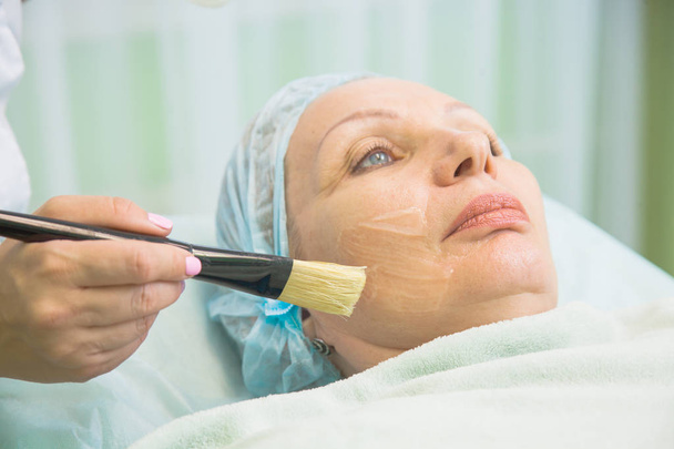 美容師は年配の女性の顔にマスクを適用します。 - 写真・画像