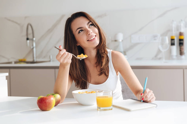 Smiling thoughtful woman having breakfast cereal and writing in notepad - Foto, imagen