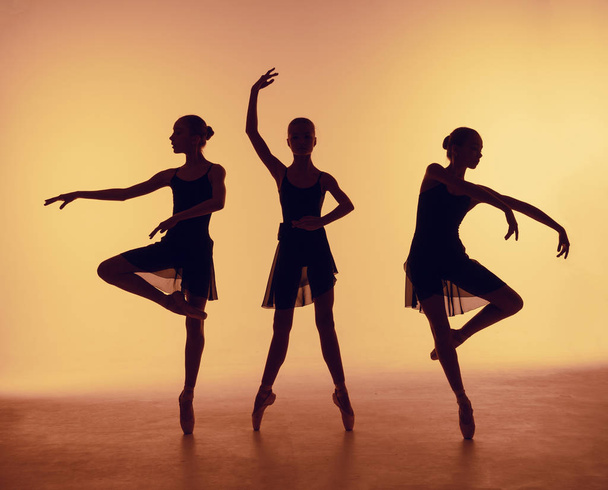 Composition from silhouettes of three young dancers in ballet poses on a orange background. - Foto, afbeelding
