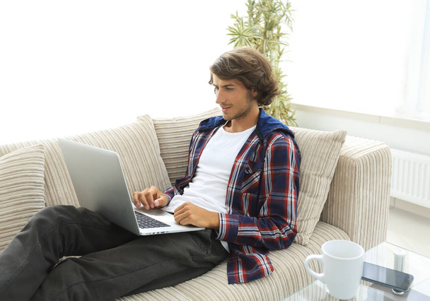 Junger Mann arbeitet am Laptop und sitzt auf Couch - Foto, Bild