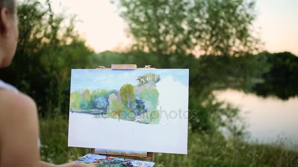 Junge Künstler malen Bild im Park mit Pinseln und Farben sitzen am romantischen See und städtischen Gebäuden im Hintergrund. Ultra-Weitwinkelblick - Filmmaterial, Video