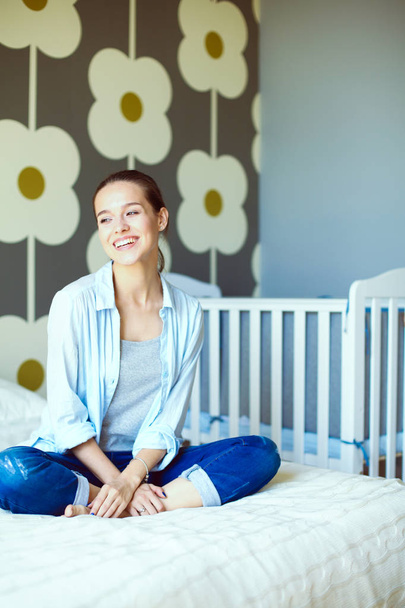 Young woman sitting on the bed near childrens cot. Young mom - Photo, Image