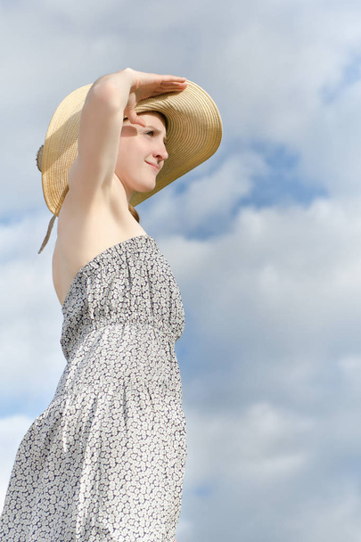Beautiful girl in hat and dress looking afar against a cloudy blue sky background - Fotó, kép