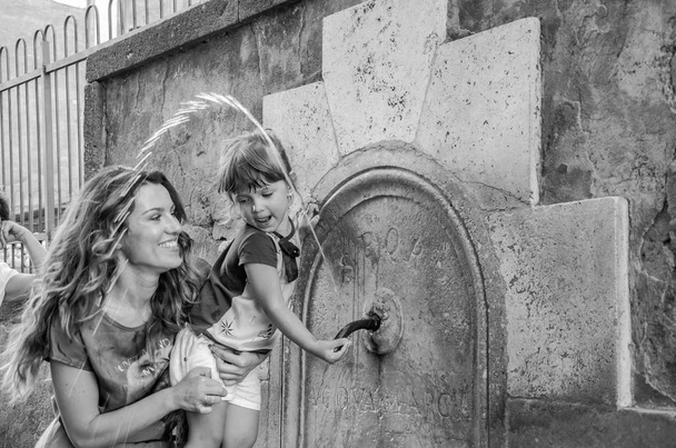 A young happy family, mother and daughter, while traveling through Italy, is played and drinks from the antique drinking fountain of the nose of Rome, on a hot summer sunny day - Photo, Image