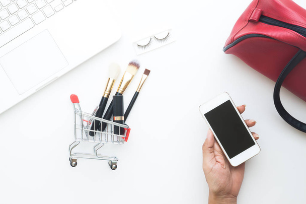 Γυναίκα χέρι κρατά κινητή συσκευή με ψώνια καλάθι και καλλυντικά είδη και laptop σε άσπρο φόντο, το Top view σε απευθείας σύνδεση εμπορική έννοια - Φωτογραφία, εικόνα
