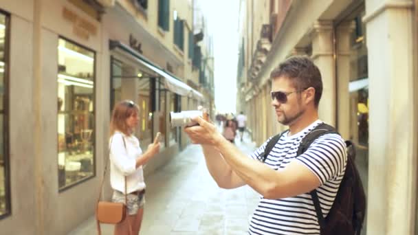 Young handsome bearded man making photos of a narrow street with his camera - Кадры, видео