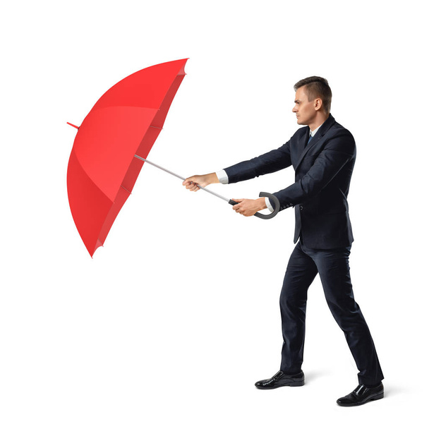 Ένας νεαρός επιχειρηματίας βρίσκεται σε λευκό φόντο και κρύβει από σκληρό περιβάλλον πίσω από μια ανοιχτή κόκκινη ομπρέλα. - Φωτογραφία, εικόνα