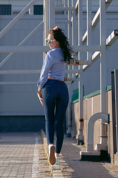 A beautiful girl with sunglasses and torn jeans and a striped shirt. Against the background of large steel structures. Artistic photography. beauty and fashion, style - Φωτογραφία, εικόνα