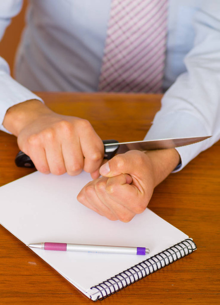 Close up of a man wants to commit suicide, using a knife in his wrist, over a notebook with a pen in a wooden table, wooden background - Photo, image