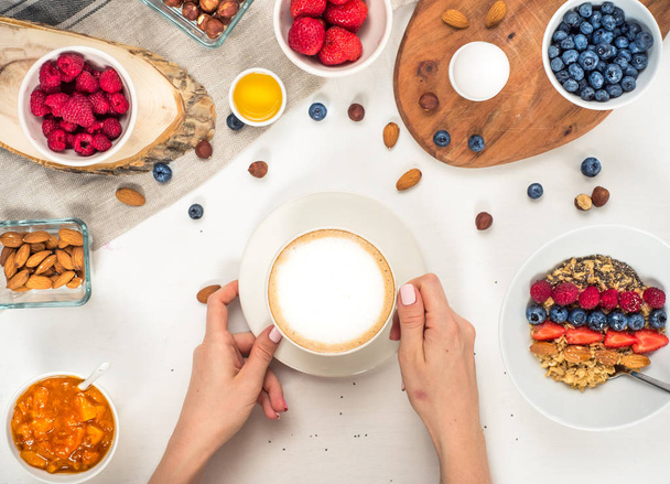 Good morning - healthy breakfast background with oatmeal coffee, berries, egg, nuts. Coffee, hands, hold, cup. White wooden food background, top view - Foto, Bild