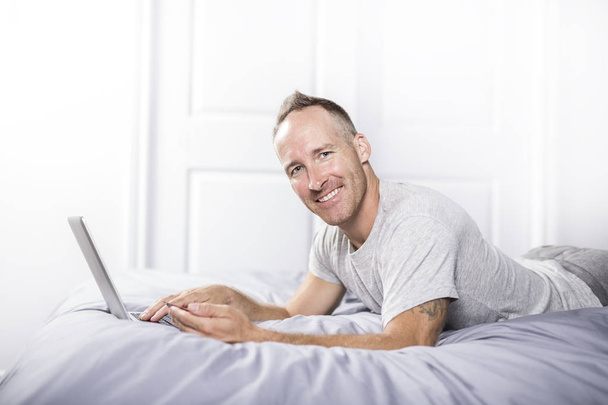 Serious casual young man using laptop in bed at home - Фото, изображение