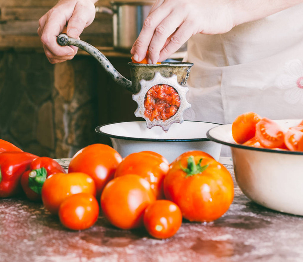 male chef grinds pieces of ripe tomatoes in an old vintage manual grinder to make homemade sauce, ketchup - Photo, image