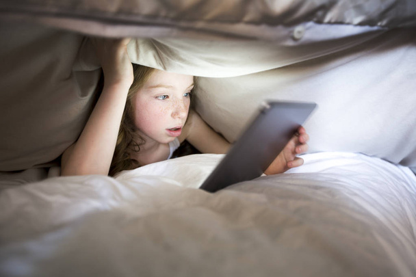 Cute little girl alone with tablet computer under blanket at night in a dark room - Foto, afbeelding
