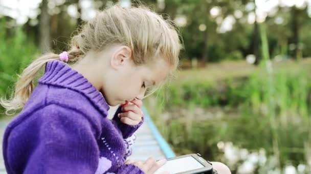 Joven hermosa chica está sentado en el puente de madera cerca del lago del bosque y está leyendo un libro electrónico
. - Metraje, vídeo