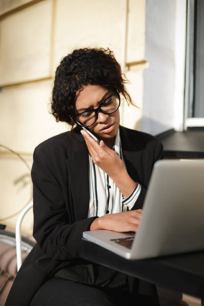 Düşünceli onu laptop bakarken onu telefonda konuşurken siyah ceketli siyah kıvırcık saçlı kadın - Fotoğraf, Görsel