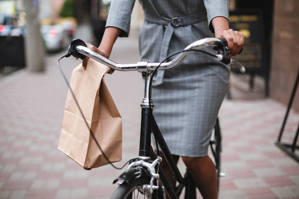 Foto da vicino del corpo della donna in abito grigio in bicicletta lungo la strada. Ritratto di giovane signora in bicicletta con sacchetto di carta in mano
 - Foto, immagini