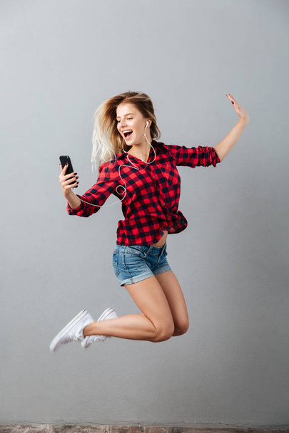 Happy young blonde woman jumping listening music by phone. - Fotoğraf, Görsel