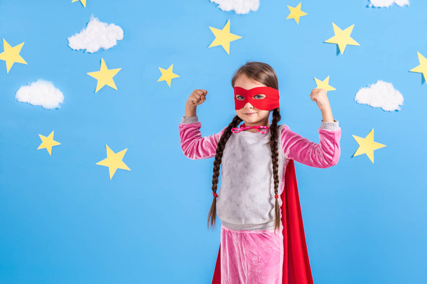 Chica rubia de seis años vestida como superhéroe divirtiéndose en casa. Niño en el fondo de la pared azul brillante
. - Foto, Imagen