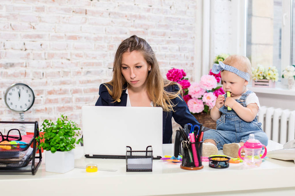 Cheerful young beautiful businesswoman looking at laptop while sitting at her working place with her little daughter - Fotografie, Obrázek
