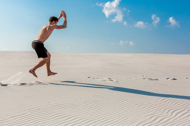 Νεαρός άνδρας άλματα στην παραλία με λευκή άμμο και φωτεινό γαλάζιο ουρανό - Φωτογραφία, εικόνα