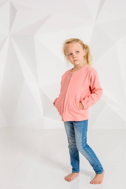 Το χαριτωμένο κοριτσάκι σε ροζ μπουφάν και τζιν, τα χέρια στις τσέπες σε λευκό φόντο - Φωτογραφία, εικόνα