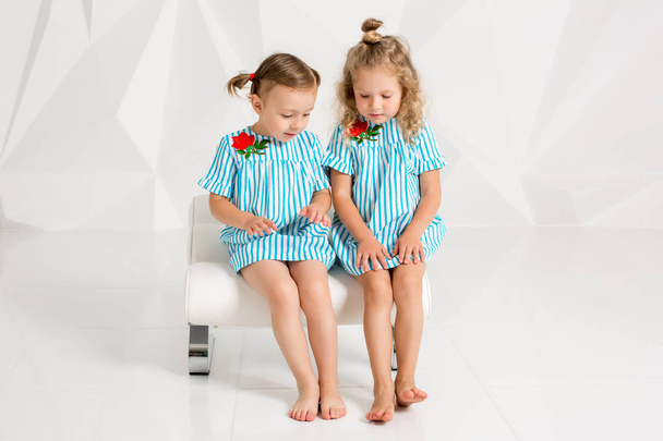 zwei kleine Freundinnen in denselben türkisfarbenen Kleidern sitzen auf einem Stuhl in einem Studio mit weißen Wänden - Foto, Bild