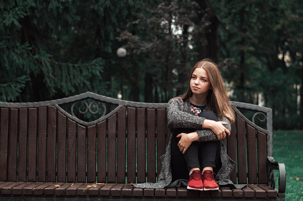 Ένα εφηβικό κορίτσι με ξανθά μαλλιά στην πλεκτή ζακέτα και μαύρο τζιν τζιν κάθεται σε ξύλινο παγκάκι στο πάρκο. Φθινόπωρο vibes - Φωτογραφία, εικόνα