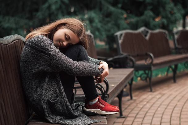 Jovem fêmea com cabelos loiros sentada no banco do parque. Uma adolescente positiva a sonhar no banco. Tempo frio de outono. Conceito de arte
 - Foto, Imagem