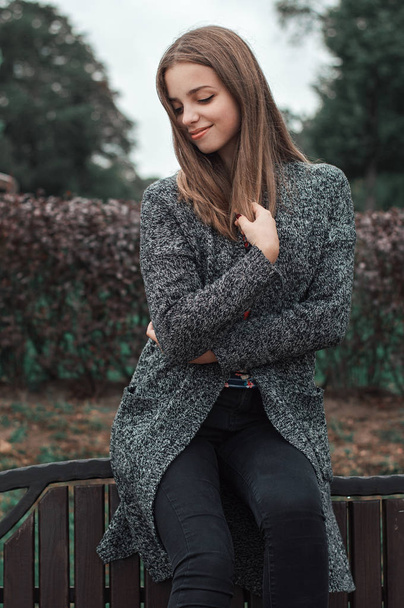 Κάθετη πορτραίτο του έφηβου κοριτσιού σε γκρι πλεκτό παλτό και μαύρο τζιν παντελόνι κάθεται στο πάρκο - Φωτογραφία, εικόνα