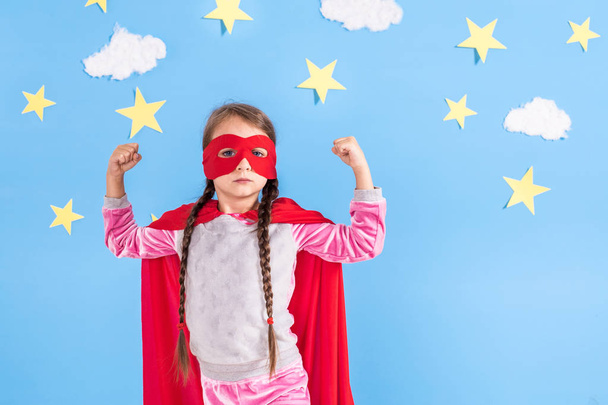 Chica rubia de seis años vestida como superhéroe divirtiéndose en casa. Niño en el fondo de la pared azul brillante
. - Foto, Imagen
