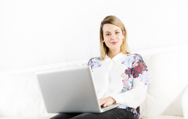 gorgeous blond woman resting on a white couch at home and using a laptop - Foto, Imagen