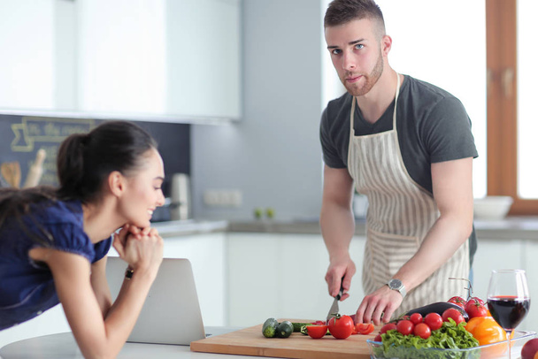 Junger Mann schneidet Gemüse und Frau steht mit Laptop in der Küche - Foto, Bild