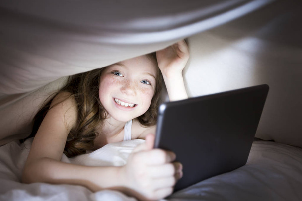 Cute little girl alone with tablet computer under blanket at night in a dark room - Valokuva, kuva