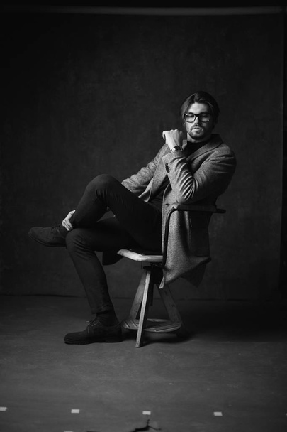 Incredibile giovane uomo sexy con bei capelli e barba e viso serio, in posa sulla sedia di legno, indossando abito con giacca grigia e orologio sulla mano, in studio grigio scuro, in bianco e nero
 - Foto, immagini