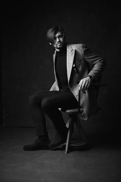 Amazing sexy young man with beautiful hair and beard and serious face, posing on the wooden chair, wearing suit with grey jacket and watch on his hand, in dark grey studio, in black and white - Foto, imagen