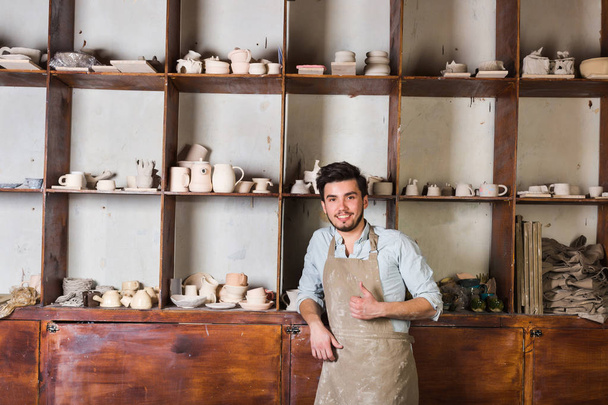 pottery, tools, ceramics art concept - happy handsome young male dressed in an apron stand with finished utensils, smiling ceramist at workplace with baked cups and jugs at background. - Foto, imagen