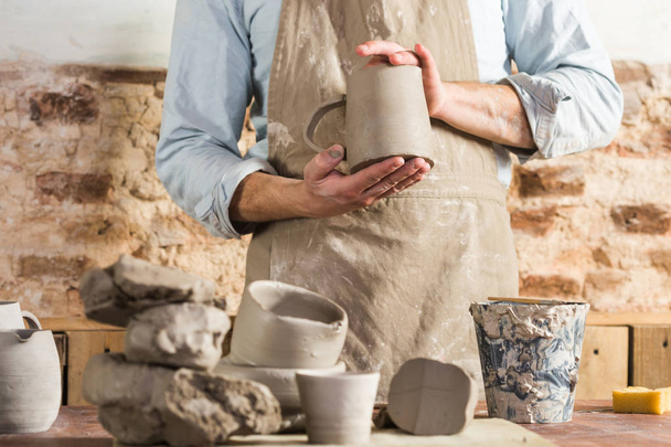 pottery, workshop, ceramics art concept - male ceramist stand at a workplace and wearing clay-stained apron, young craftsmans hands holding unbaked jug, workplace with fireclay and unfinished cups. - Photo, Image
