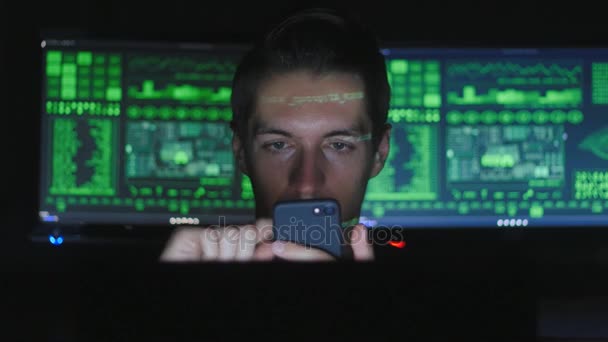Programador Hacker utiliza un teléfono inteligente, mientras que los caracteres de código verde reflejan en su cara en una habitación oscura oficina
. - Metraje, vídeo