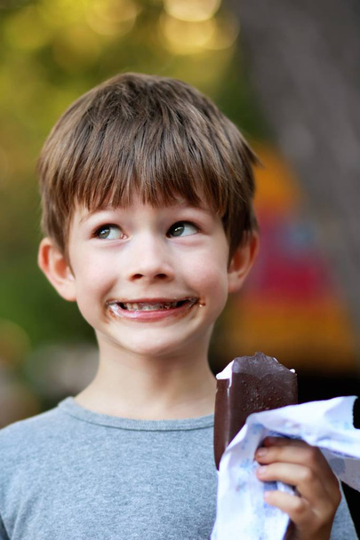 Happy boy in grey t shirt making faces while eating ice cream outdoor in the park. Close up funny facial expression, blurred - Photo, Image