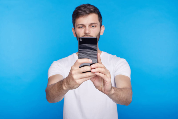 Χαρούμενος νεαρός hipster Καυκάσιος κρατώντας το Γενόσημο κινητό τηλέφωνο και χαμογελώντας γενικές γραμμές κατά τη λήψη selfie. Πάντα σε επικοινωνία. Επιλεκτική εστίαση στο χέρι με gadget. Άνθρωποι, σύγχρονο τρόπο ζωής και την τεχνολογία. - Φωτογραφία, εικόνα