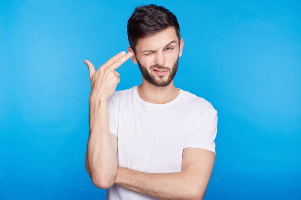 Isolated portrait of European young international student tired from passing exams, shooting himself with finger gun gesture against blank blue wall background. Human face expressions and emotions. - Photo, Image